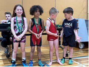Young children wearing medals after a wrestling event, standing together on a gym floor.
